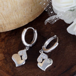 Load image into Gallery viewer, Silver heart-shaped earrings on a wooden surface with white flowers.
