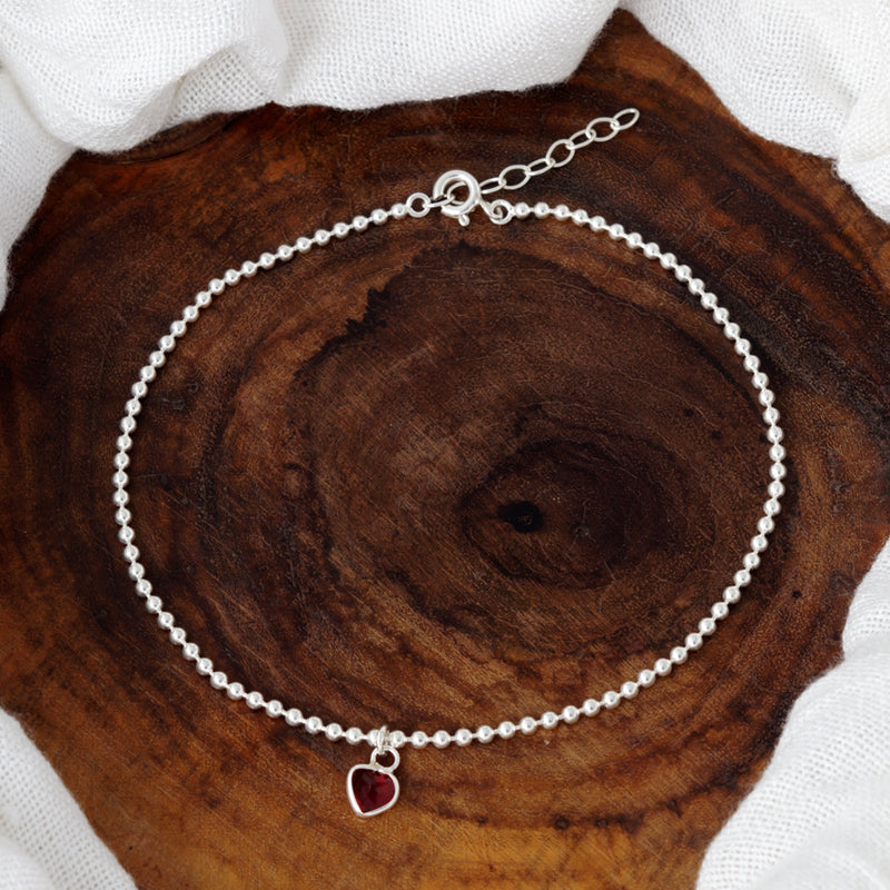 Silver beaded anklet with a red heart charm on a purple and blue background