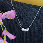 Load image into Gallery viewer, Silver necklace with floral pendant on a textured black surface with pink flowers.
