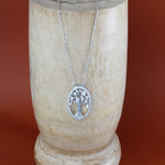 Load image into Gallery viewer, Silver necklace with a tree of life pendant on a stone stand against a red background
