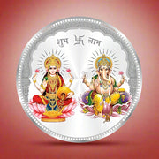 A hallmarked round silver coin with the depiction of two figures, likely to represent Laxmi and Ganesh, surrounded by intricate designs and floral motifs.